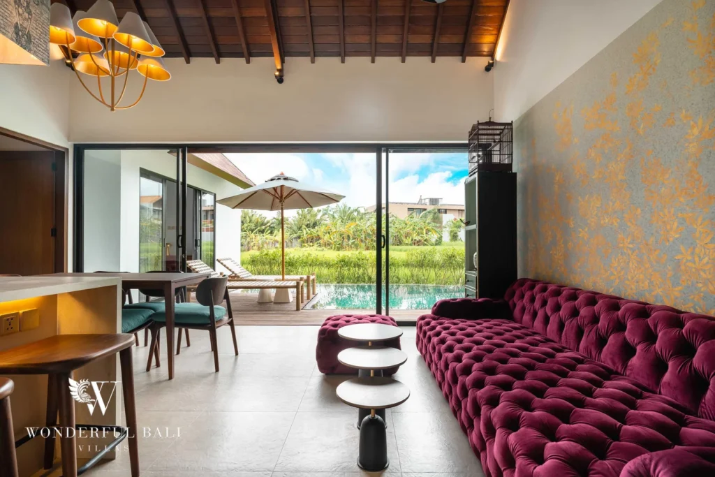 A magenta velvet sofa and dining area under a vaulted timber ceiling opening through glass doors to the poolside terrace and rice fields at a 4-bedroom villa in Canggu, Bali