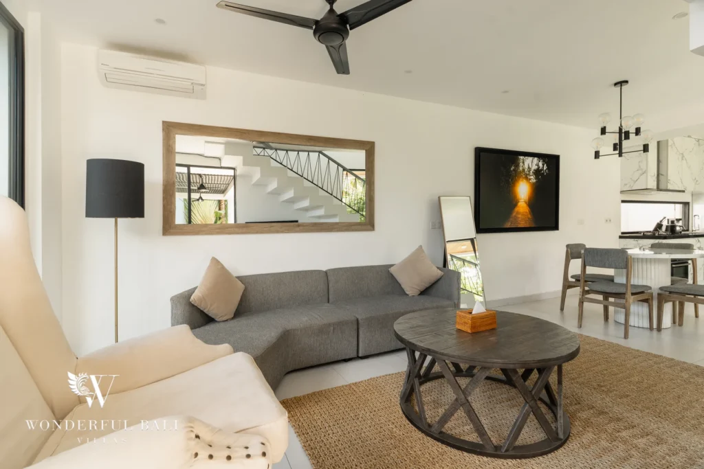 An enclosed living room with a grey sofa, cream armchair, round coffee table, ceiling fan, air conditioning and a framed mirror beside the dining area at a 3-bedroom villa in Berawa, Bali