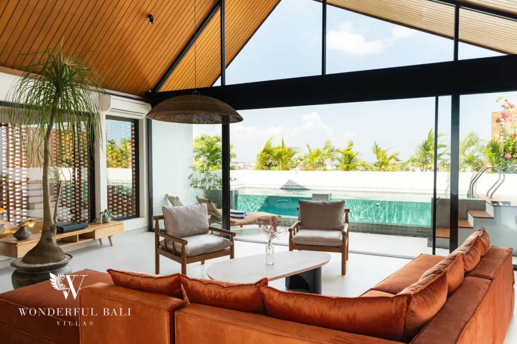 An enclosed living room with burnt-orange sofas and timber chairs under a vaulted wood ceiling, opening through glass walls to a pool and rice-field view at a 5-bedroom villa in Batu Bolong, Canggu, Bali