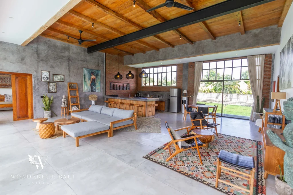 An open loft-style living room with concrete and brick walls, timber ceiling, ceiling fans and mid-century chairs on a patterned rug at a 3-bedroom villa in Seminyak, Bali