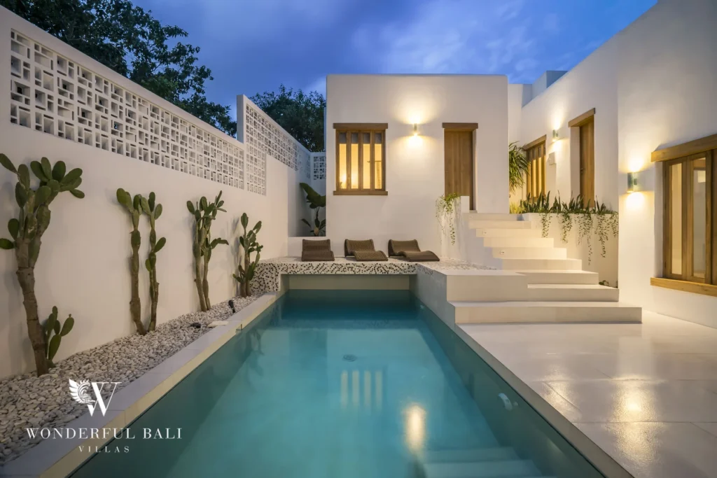 A slim turquoise pool lined with potted cacti and pebble beds reflecting warm lights from the white villa walls in the evening at a 3-bedroom villa in Bingin, Uluwatu, Bali