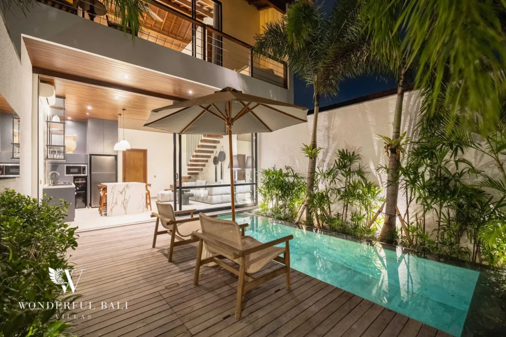 The pool and wooden deck lit at night with an umbrella, chairs and a glowing interior below an upstairs balcony at a 2-bedroom villa in Canggu, Bali