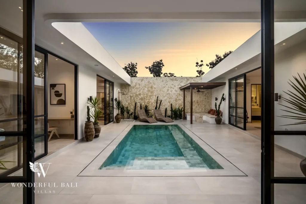 A private pool framed by a natural stone wall, sun loungers and potted plants at dusk viewed from inside at a 3-bedroom villa in Bingin, Uluwatu, Bali