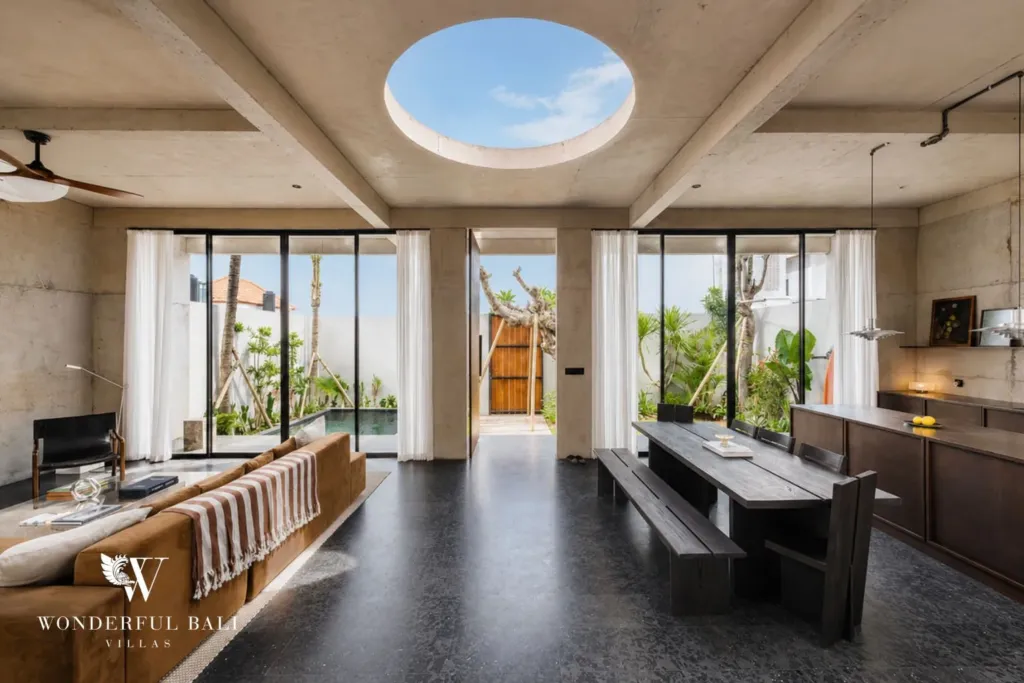 Open-plan living and dining space with a round ceiling skylight, timber dining bench and sofa facing the pool through steel-framed glass at a 2-bedroom villa in Seseh, Bali