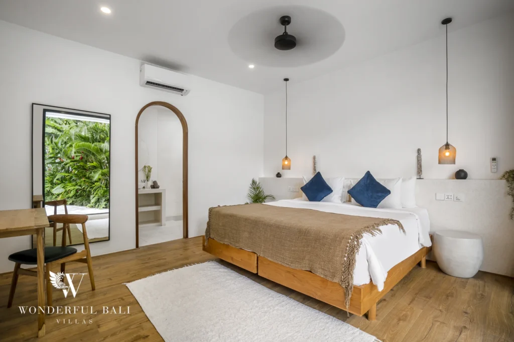 An en-suite bedroom with a wooden bed, blue cushions, arched mirror and garden view through a large window at a 3-bedroom villa in Bingin, Uluwatu, Bali