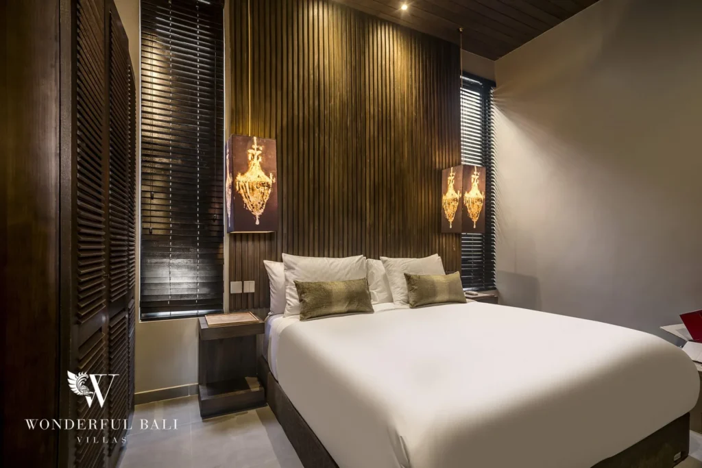 King bed against a wood-slat wall lit by ornate chandelier sconces with shuttered windows at a 2-bedroom villa in Pererenan, Bali
