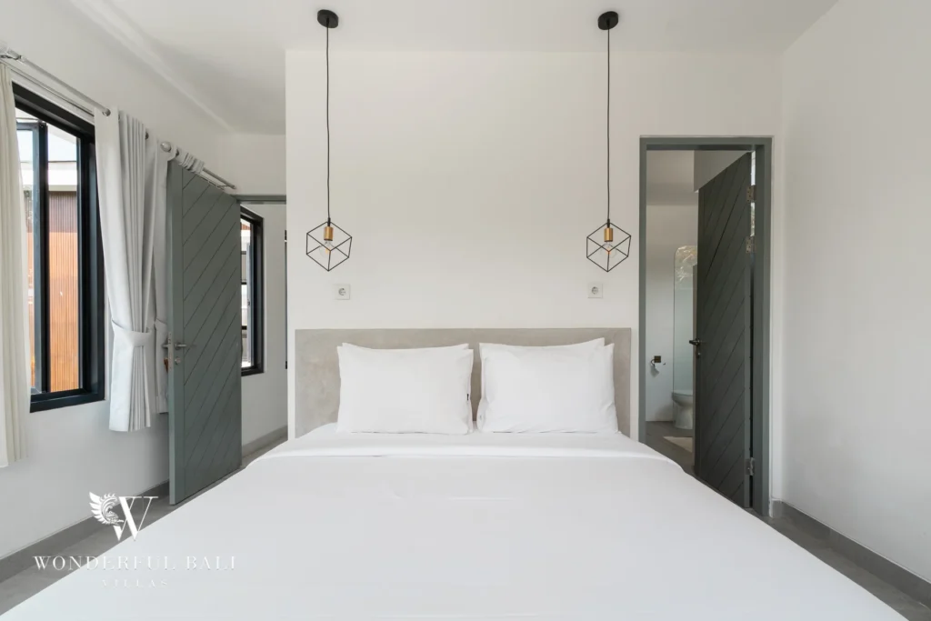 A bedroom with a white double bed, geometric pendant lights and an open door leading to an en-suite bathroom at a 3-bedroom villa in Berawa, Bali