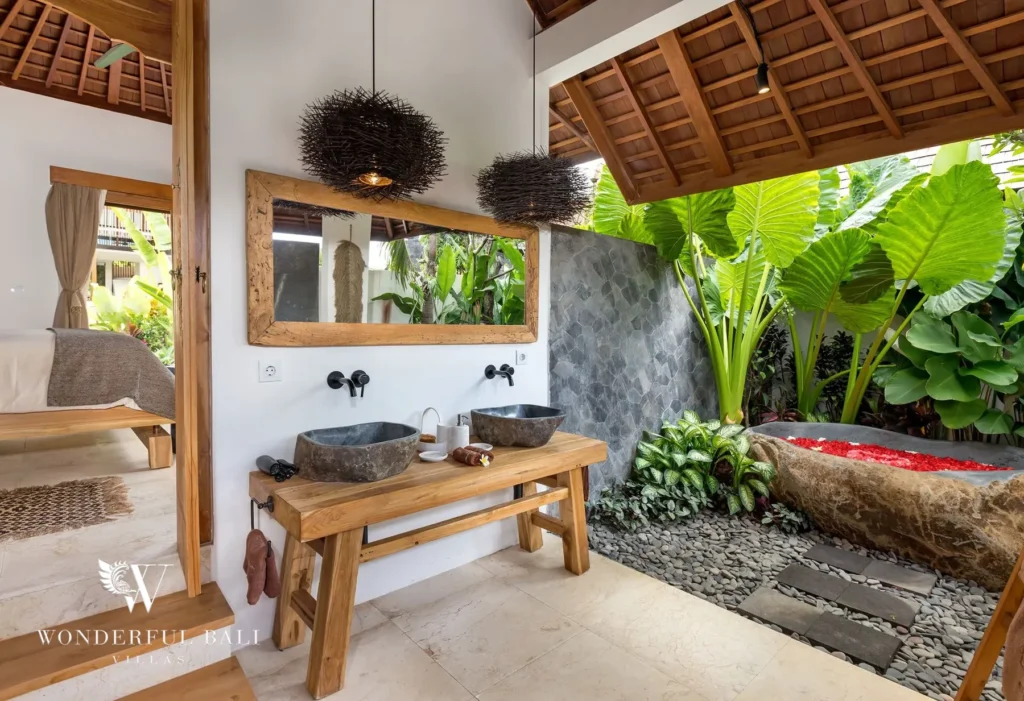 An open-air en-suite bathroom with twin stone basins on a wood vanity and a natural stone bathtub among tropical plants at a 4-bedroom villa in Canggu, Bali