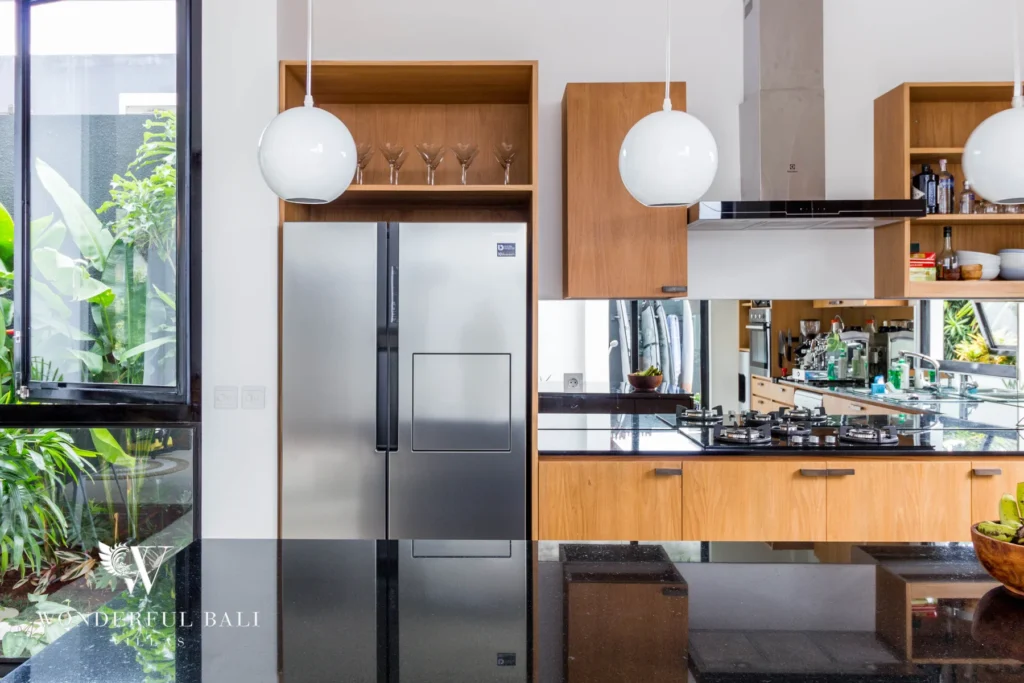A kitchen with a stainless steel fridge, gas hob, timber cabinetry and a black-framed window onto tropical greenery at a 4-bedroom villa in Batu Bolong, Canggu, Bali