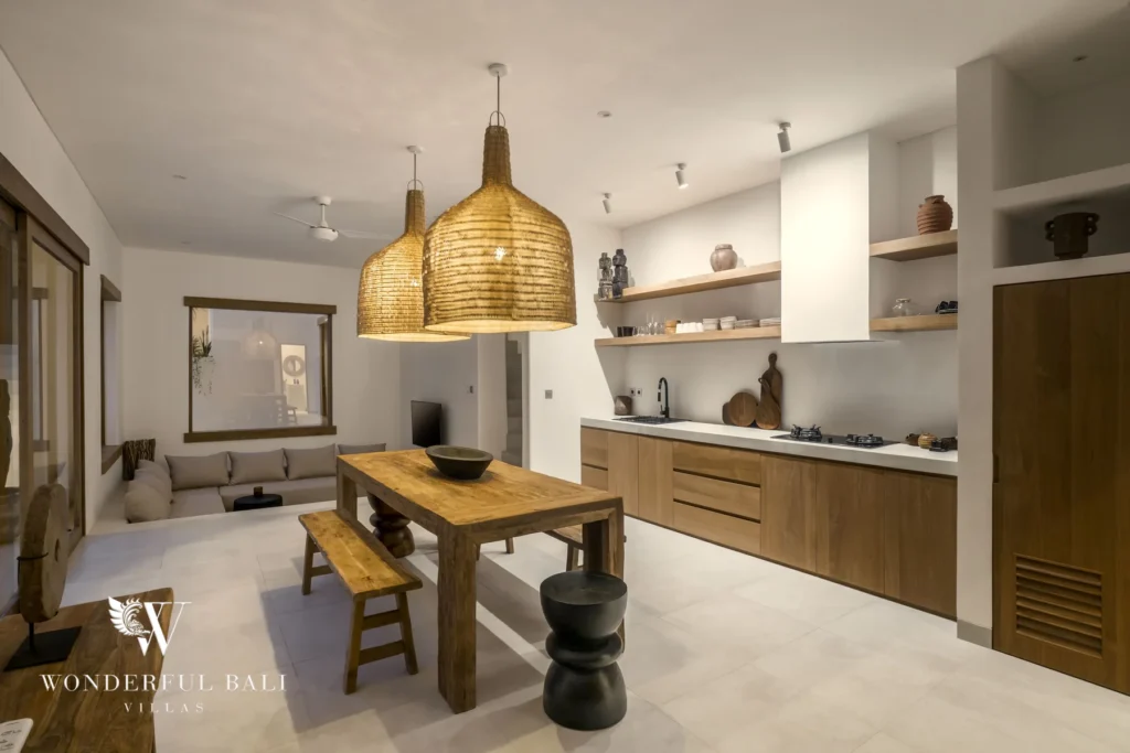 A wooden dining table with a bench under woven pendant lamps beside a fitted timber kitchen and a cushioned sofa nook at a 3-bedroom villa in Bingin, Uluwatu, Bali