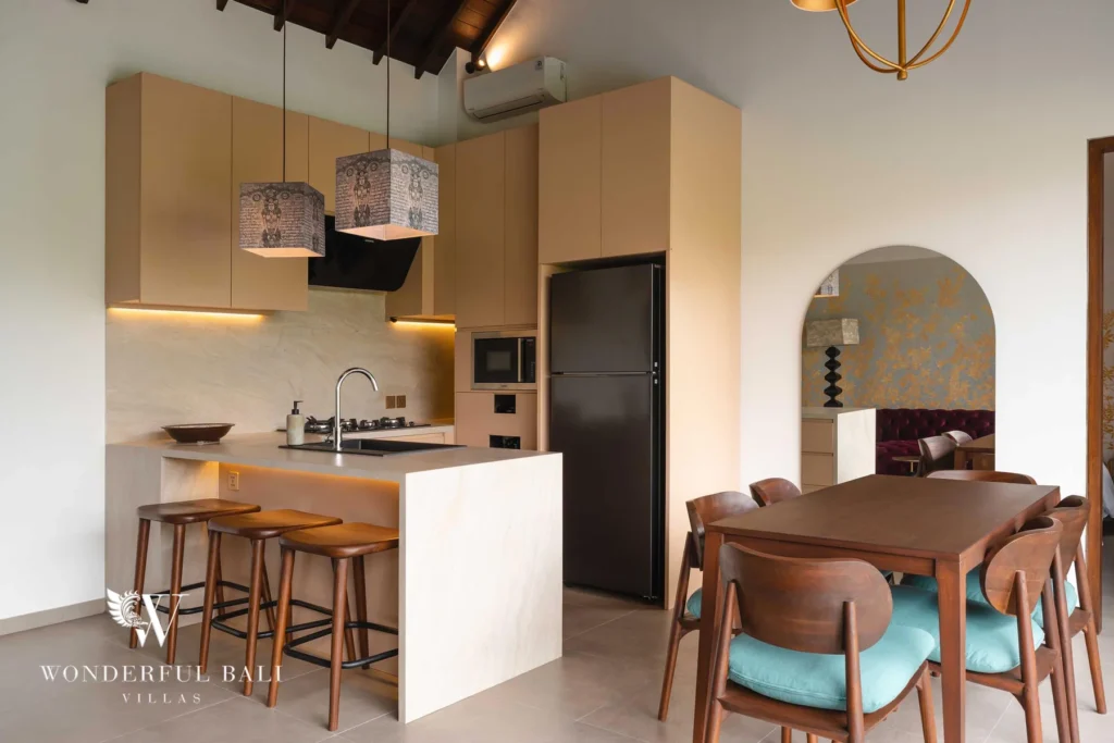 An open-plan kitchen with pale cabinetry, patterned pendant lamps and a stone island with wooden stools next to a wooden dining table with teal chairs at a 4-bedroom villa in Canggu, Bali