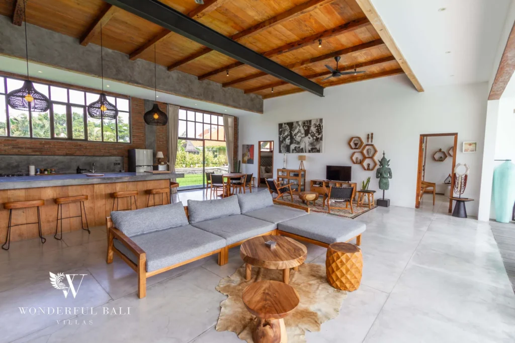 A warehouse-style living space with grey sofas, a bar counter with stools, timber ceiling and steel-framed windows opening to greenery at a 3-bedroom villa in Seminyak, Bali