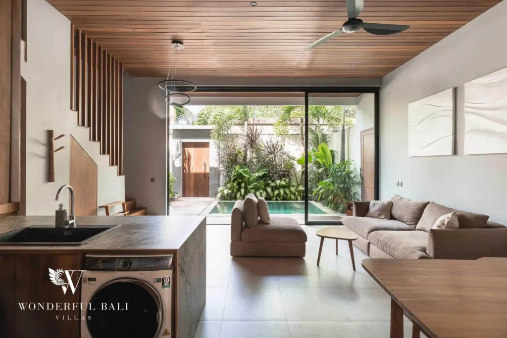 2BR villa for rent in Pererenan — living room, an open-plan kitchen with a stone island and built-in washing machine flowing to a sofa lounge and pool view under a timber ceiling at a 2-bedroom villa