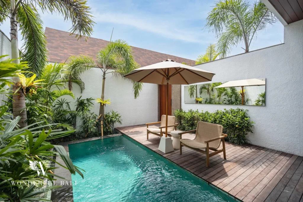 A private pool bordered by a wooden deck with two chairs, an umbrella and lush palms at a 2-bedroom villa in Canggu, Bali