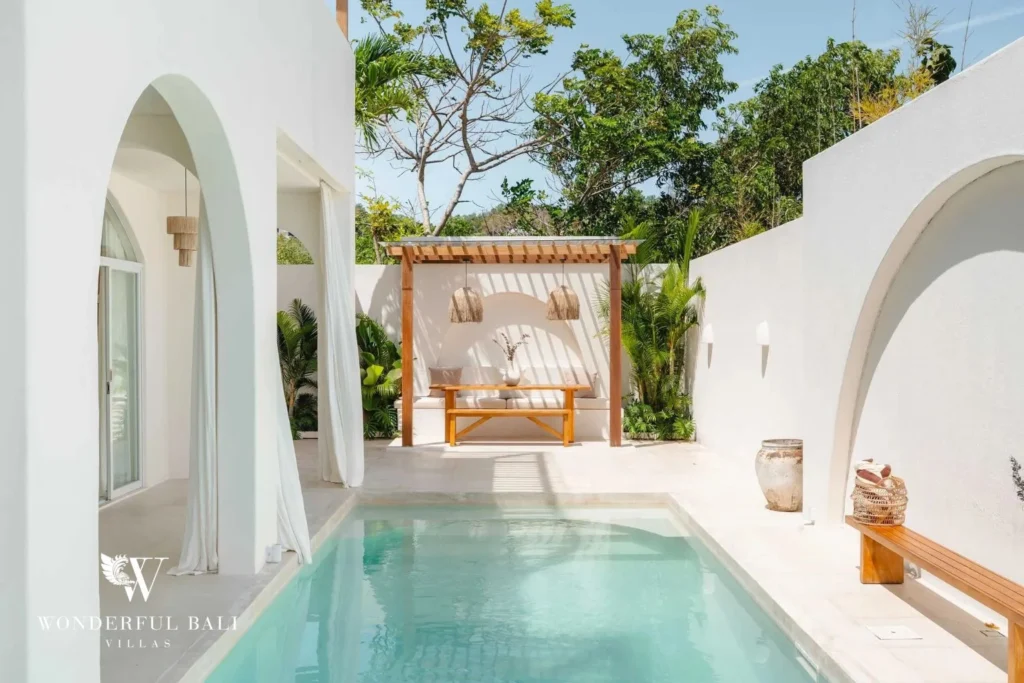 The pool with a wooden pergola daybed and dining bench surrounded by tropical greenery and white walls, at a 2-bedroom villa in Bingin, Uluwatu, Bali