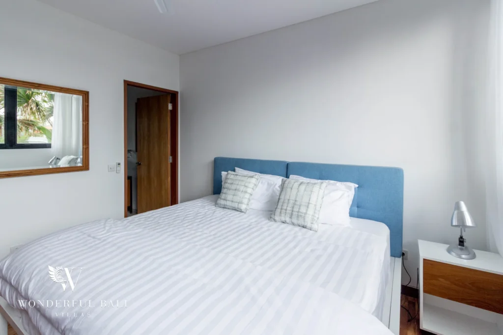 A bedroom with a blue upholstered headboard, striped white bedding and a wall mirror beside a timber door at a 4-bedroom villa in Batu Bolong, Canggu, Bali