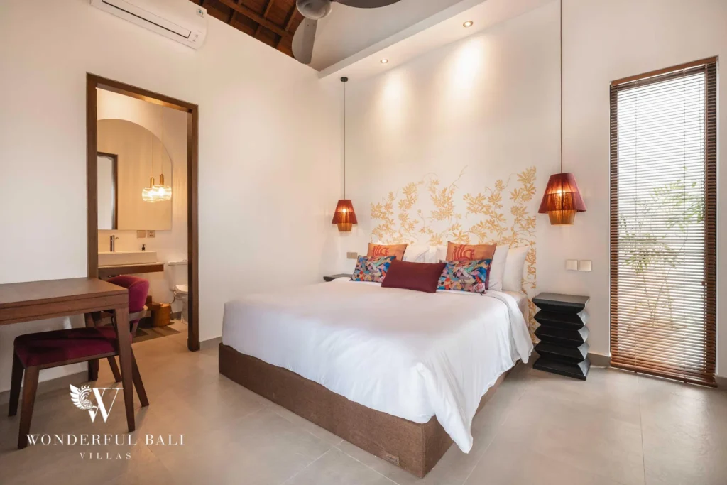 A bedroom with a floral gold headboard, colourful cushions and red pendant lamps beside a wooden desk and an en-suite bathroom doorway at a 4-bedroom villa in Canggu, Bali