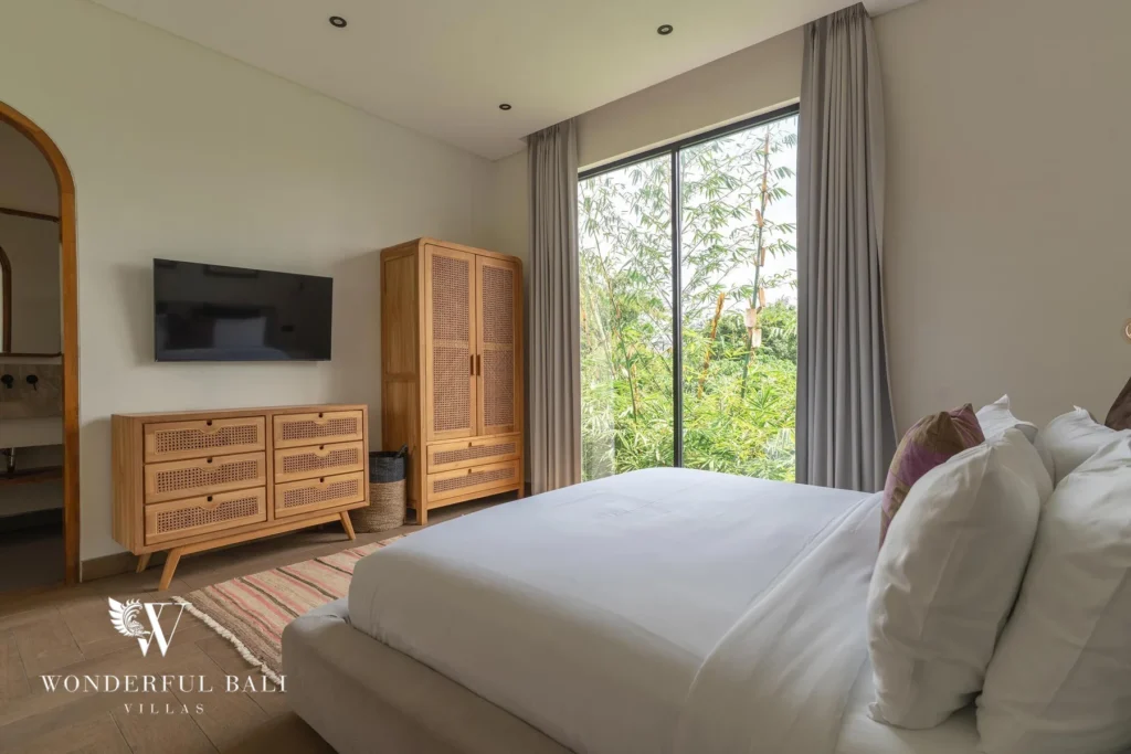 A bedroom with a wall-mounted smart TV, rattan wardrobe and a floor-to-ceiling window onto tropical greenery at a 2-bedroom villa in Canggu, Bali