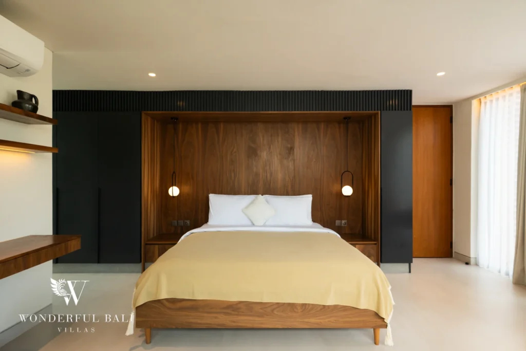 A spacious bedroom with a walnut wood headboard wall, dark cabinetry, twin globe pendant lights and a yellow bed throw at a 5-bedroom villa in Batu Bolong, Canggu, Bali