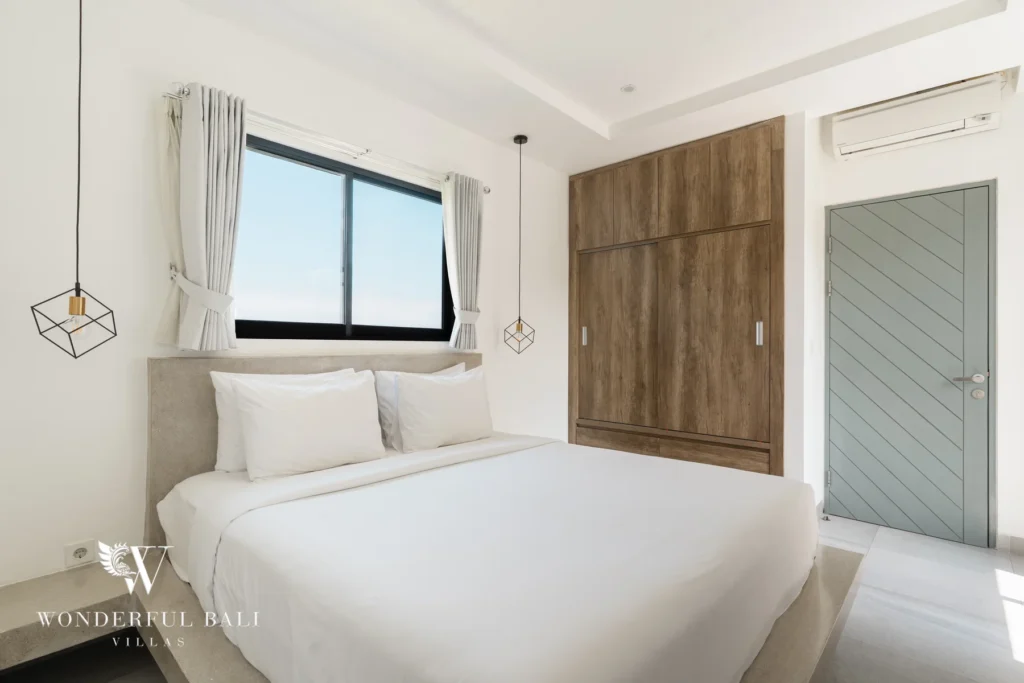 A bright bedroom with a double bed, window, large wooden wardrobe, air conditioning and a grey timber door at a 3-bedroom villa in Berawa, Bali