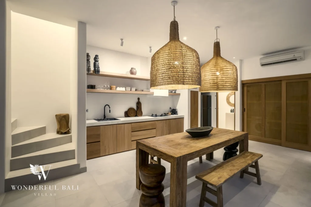A rustic wooden dining table and stools sit under oversized woven pendants beside a timber kitchen counter and a short flight of steps at a 3-bedroom villa in Bingin, Uluwatu, Bali