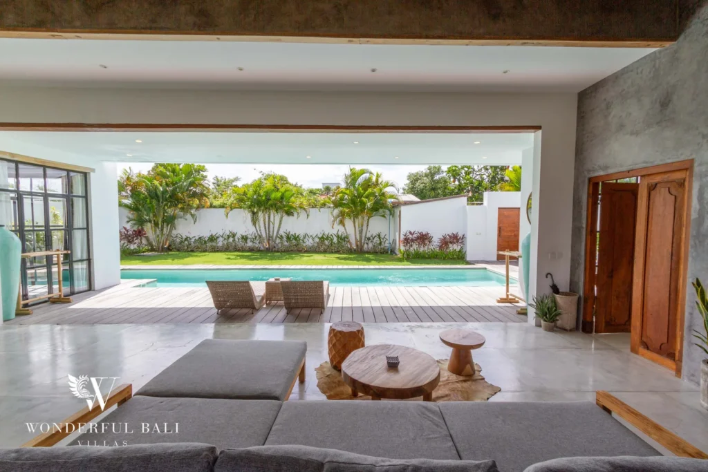 The open-sided lounge looks out through wide sliding doors onto the pool, timber deck and palm-lined garden at a 3-bedroom villa in Seminyak, Bali