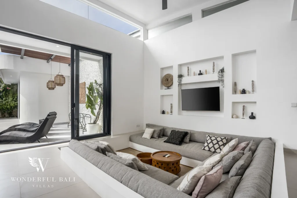 Cozy living room with a large grey sunken sofa, wall-mounted TV and niche shelving beside sliding doors to a lounger terrace at a 3-bedroom villa in Bingin, Uluwatu, Bali