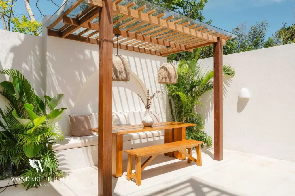 A shaded outdoor nook with a timber pergola, built-in cushioned bench, wooden dining table and lush plants, at a 2-bedroom villa in Bingin, Uluwatu, Bali