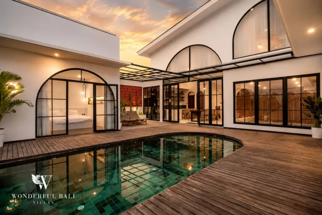 The green mosaic pool and wooden deck glow at dusk beneath arched steel-frame doors and a warm sunset sky at a 3-bedroom villa in Pererenan, Bali