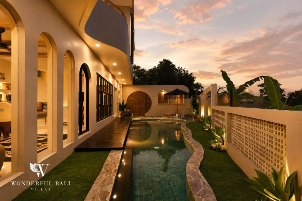 The illuminated pool and villa facade glow at dusk beside a wooden deck and garden at a 3-bedroom villa in Pererenan, Bali