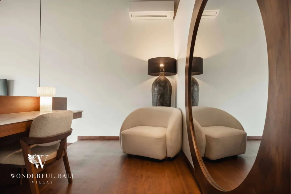A reading nook with a cream armchair, dark table lamp, desk and a large round wooden mirror at a 2-bedroom villa in Canggu, Bali