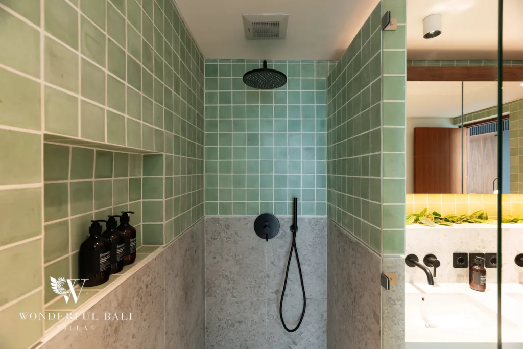 An en-suite bathroom with a green-tiled walk-in rain shower, grey stone lower walls, black fixtures and a mirrored vanity reflection at a 5-bedroom villa in Batu Bolong, Canggu, Bali