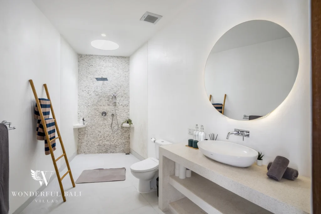 Bright bathroom with a round backlit mirror, stone vanity, walk-in mosaic-tiled shower and a bamboo towel ladder at a 3-bedroom villa in Bingin, Uluwatu, Bali