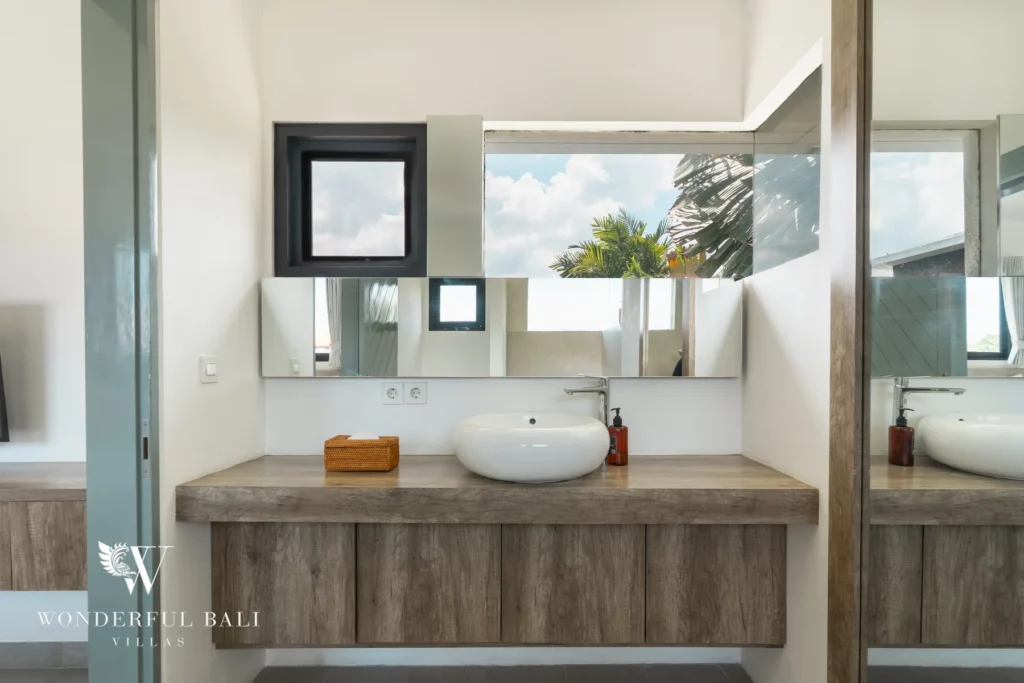 A bathroom with a wooden floating vanity, round vessel basin, wide mirror and square window framing palm trees at a 3-bedroom villa in Berawa, Bali