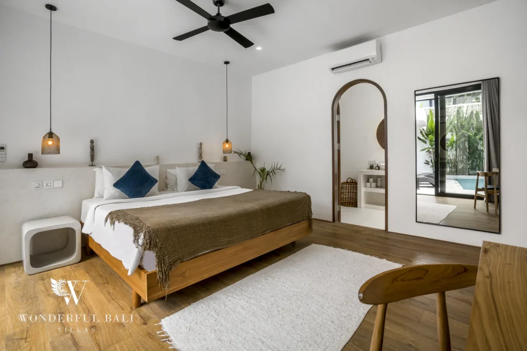 A spacious en-suite bedroom with a wooden bed, blue cushions, arched dressing nook and pool-view window at a 3-bedroom villa in Bingin, Uluwatu, Bali