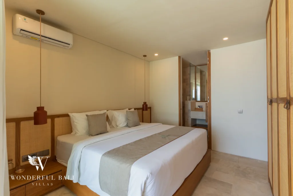 A neutral-toned bedroom with a double bed, rattan pendant lights and an ensuite bathroom visible through the doorway at a 3-bedroom villa in Bingin, Uluwatu, Bali