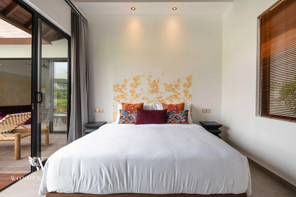 A king bed with a gold floral headboard and colourful cushions beside sliding glass doors opening to a terrace with loungers and rice-field views at a 4-bedroom villa in Canggu, Bali