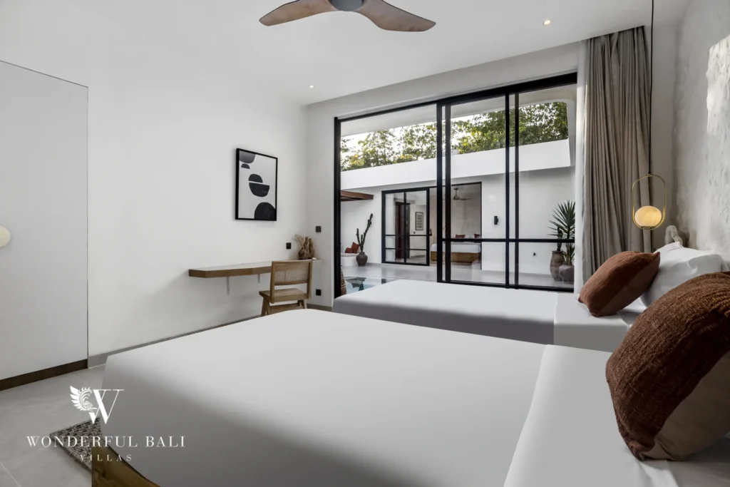A twin bedroom with two beds, a wall-mounted desk and glass doors overlooking the pool courtyard at a 3-bedroom villa in Bingin, Uluwatu, Bali
