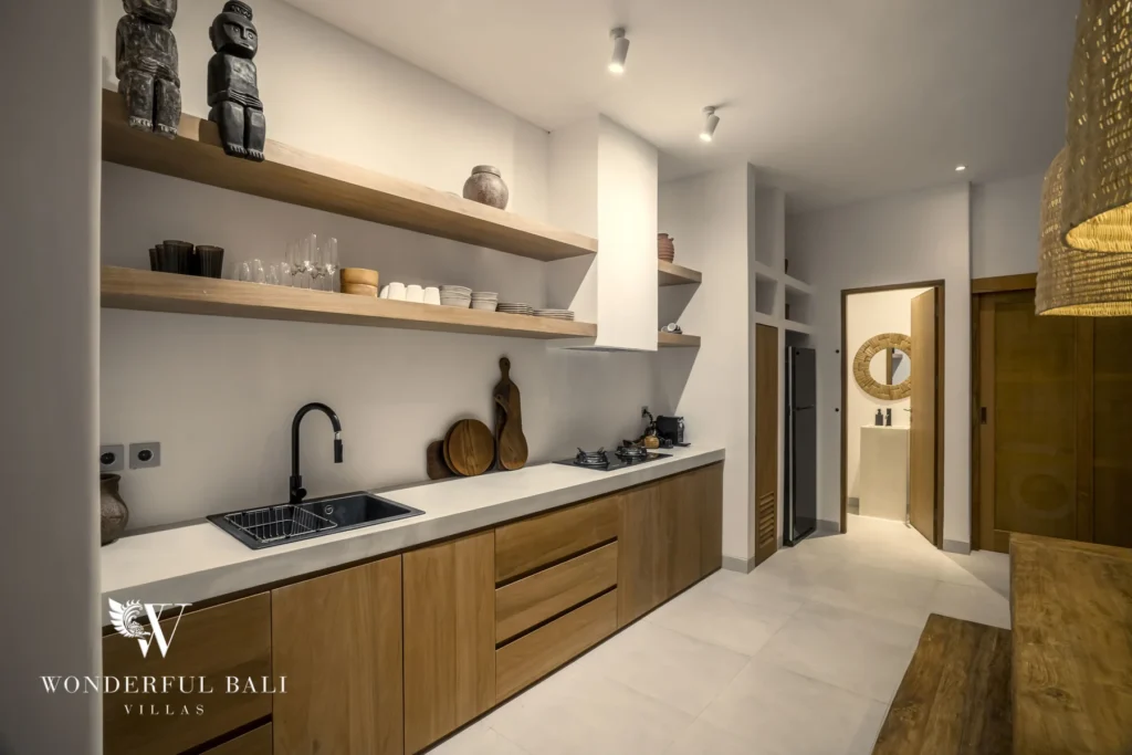 A galley kitchen with wooden cabinetry, open shelves, a black sink and gas hob leading to a small bathroom at a 3-bedroom villa in Bingin, Uluwatu, Bali