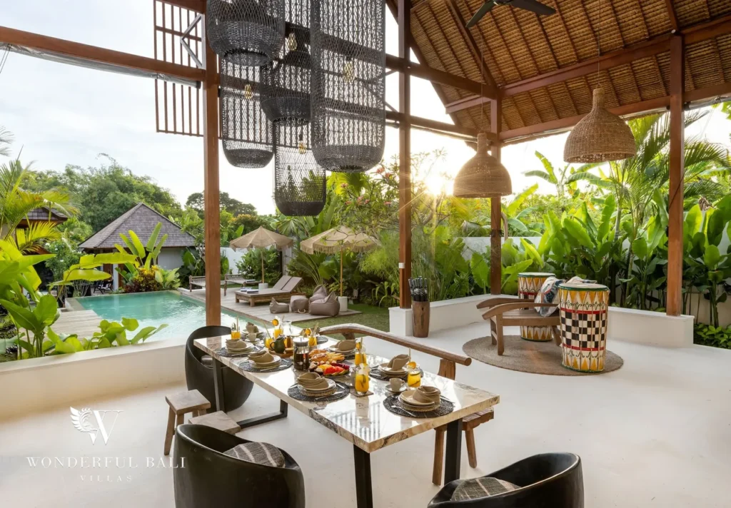 An open pavilion dining area with a marble table, black chairs and woven pendant lamps looks out to the pool, sun loungers and garden at a 4-bedroom villa in Canggu, Bali