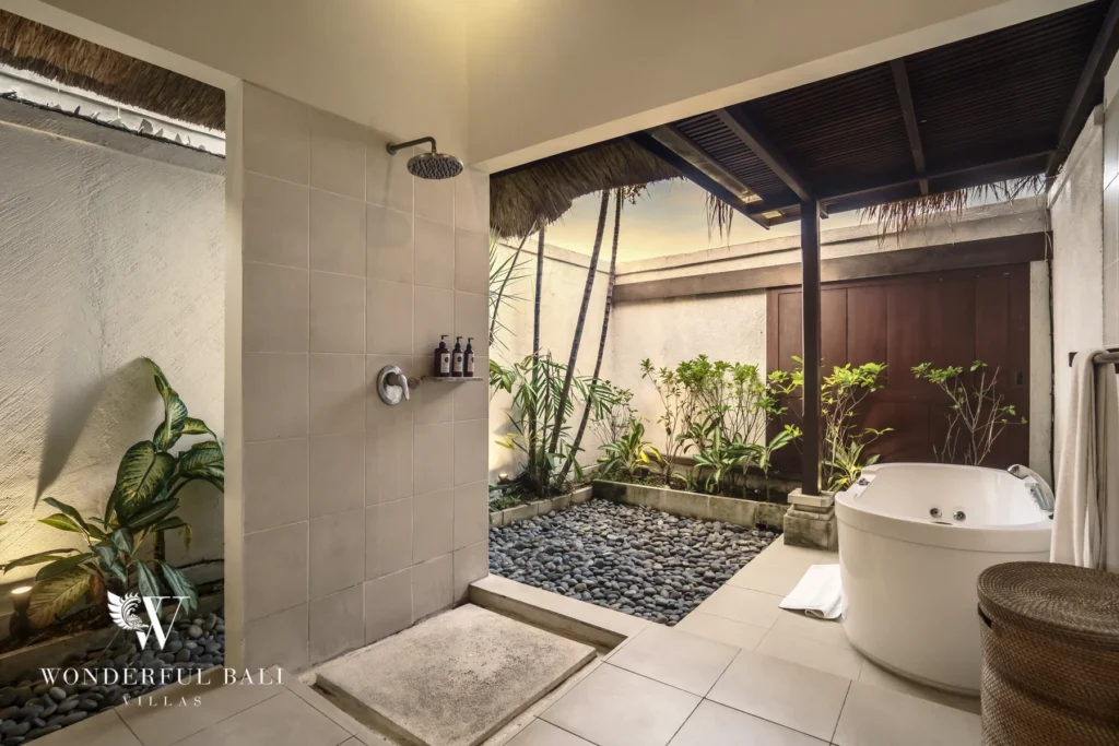 A semi-open bathroom with a freestanding bathtub, rain shower and tropical plants over pebble stones at a 4-bedroom villa in Seminyak, Bali
