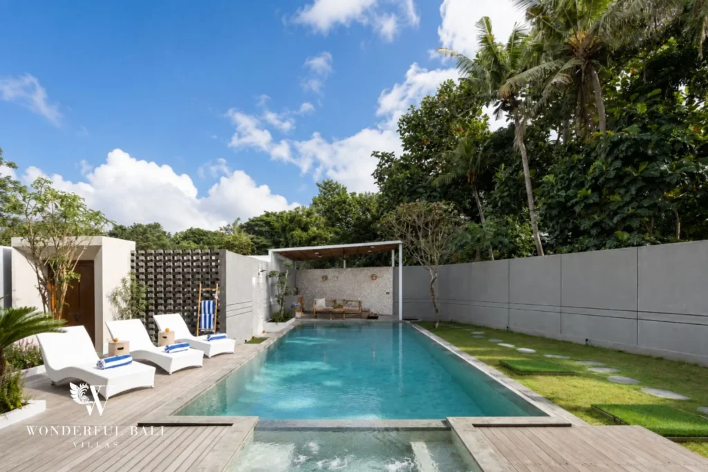 A long pool flanked by white sun loungers, a timber deck and a covered lounge backed by tropical greenery at a 4-bedroom villa in Pererenan, Bali