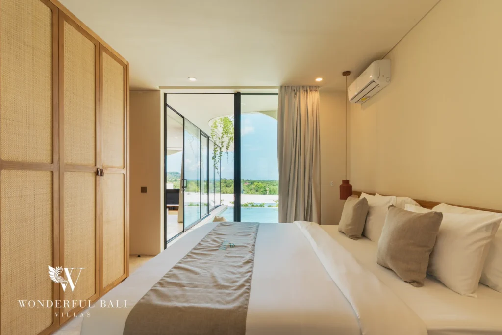 A bedroom with rattan wardrobes and a floor-to-ceiling glass wall opening to a pool and panoramic landscape at a 3-bedroom villa in Bingin, Uluwatu, Bali