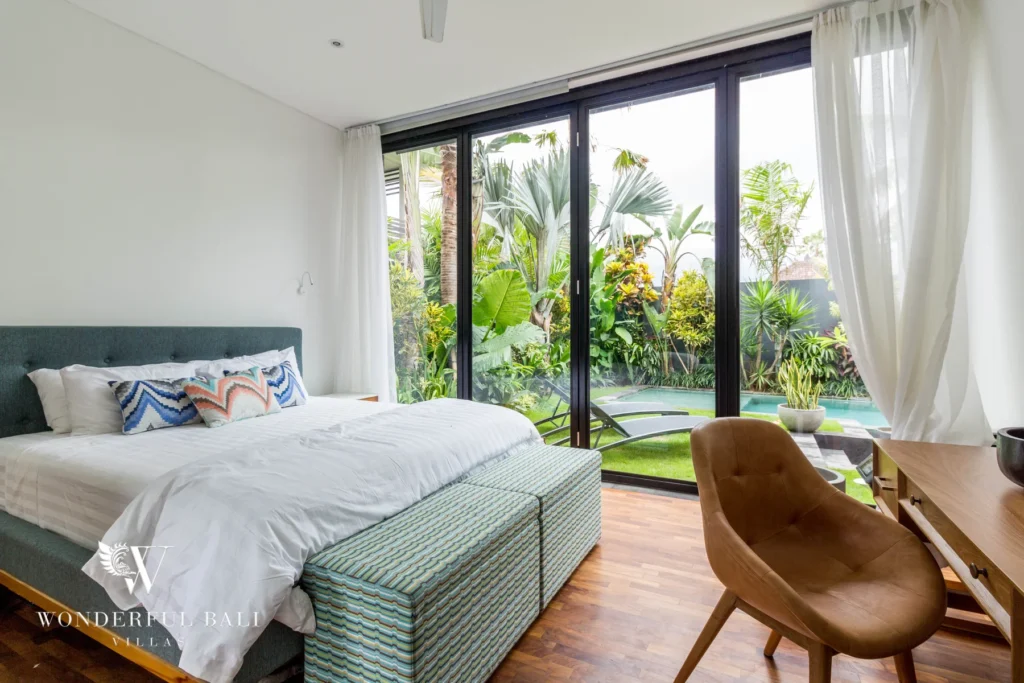 A bedroom with retractable glass doors opening to the pool and lush garden, a tan lounge chair and desk at a 4-bedroom villa in Batu Bolong, Canggu, Bali