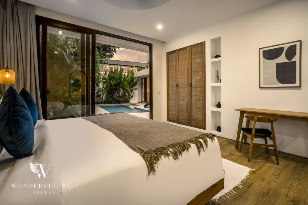 An en-suite bedroom with a white bed, wooden wardrobe and sliding doors opening onto the pool at dusk at a 3-bedroom villa in Bingin, Uluwatu, Bali