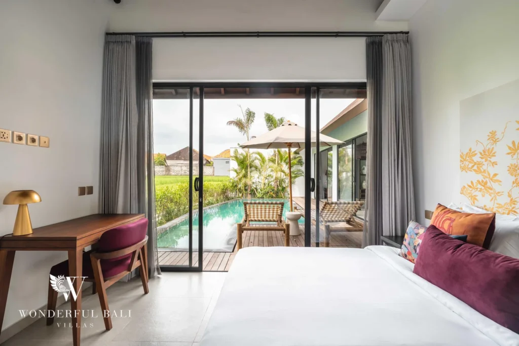 A bedroom with a wooden desk and magenta chair by a window, the bed beside sliding doors framing the pool, umbrella and rice fields at a 4-bedroom villa in Canggu, Bali