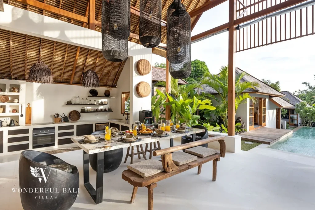 An open pavilion holding a stone dining table with benches and an open kitchen with white cabinetry looks out to the pool and garden bungalows at a 4-bedroom villa in Canggu, Bali