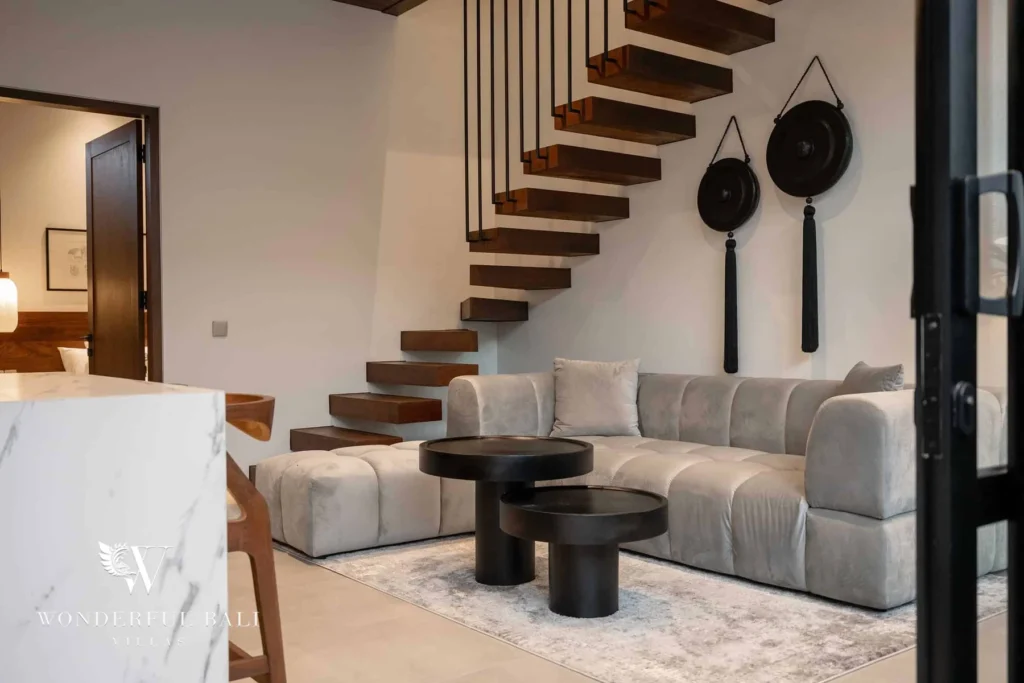 A grey sectional sofa with black round coffee tables beneath a floating staircase and wall-mounted gongs at a 2-bedroom villa in Canggu, Bali