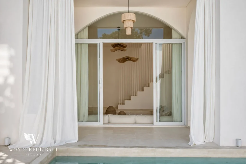 A view from the pool through tall glass doors into an arched interior with a pale staircase and sheer curtains, at a 2-bedroom villa in Bingin, Uluwatu, Bali