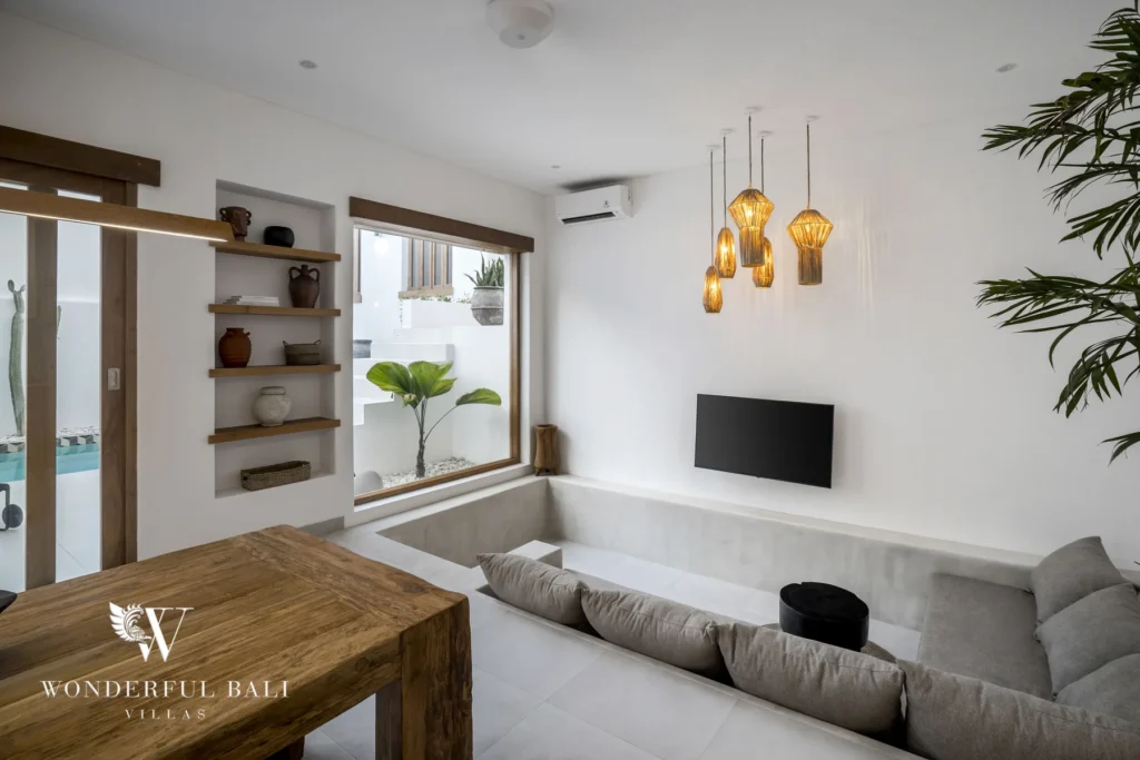A sunken grey sofa lounge with amber pendant lights, a wall-mounted TV, a wooden table and a large picture window overlooking the pool at a 3-bedroom villa in Bingin, Uluwatu, Bali